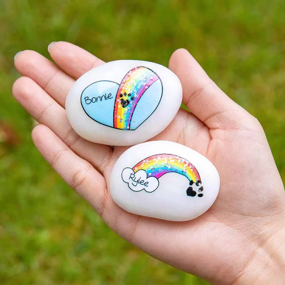 Two engraved rocks with a rainbow and pet name Bonnie held in a hand, with a sequin finish.