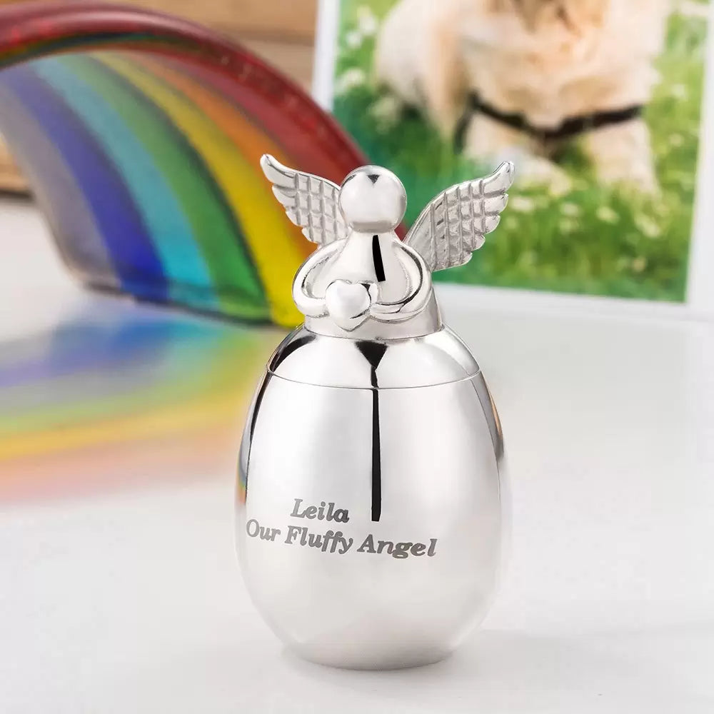 A silver stainless steel heart-shaped pet ashes urn with an angel design, personalized with the name 'Leila' and the phrase 'our fluffy angel' on the front.