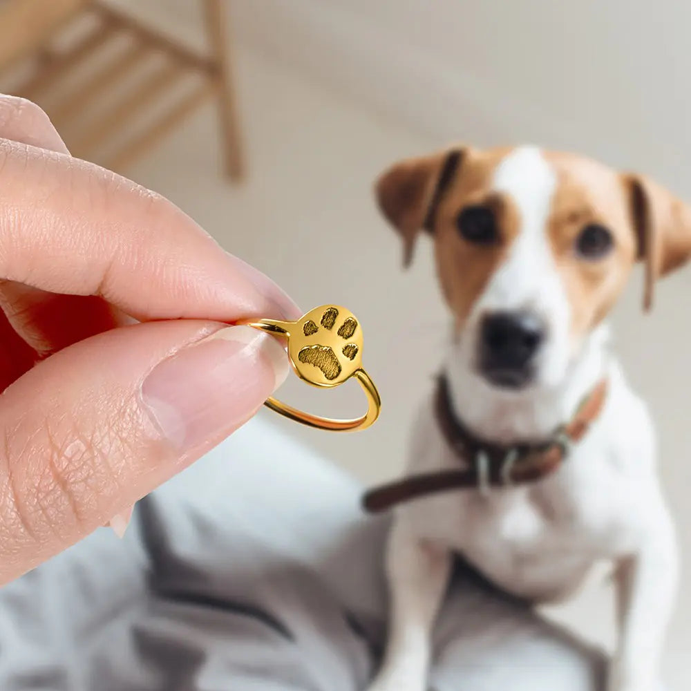 Custom pet paw print ring held by hand with dog in background, personalized ring engraved from pet paw print