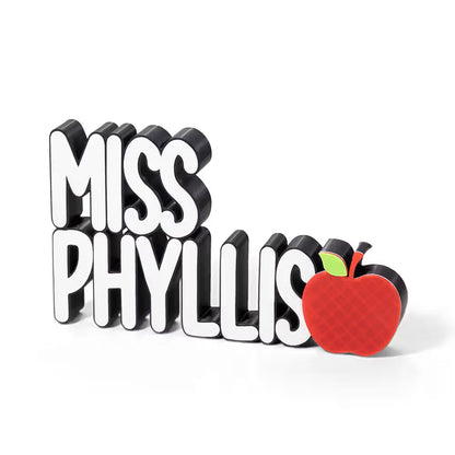 A custom 3D block-style sign reading 'MISS PHYLLIS' in white letters with black sides, plus a red apple with a green leaf on the right, shown on a clean white background.