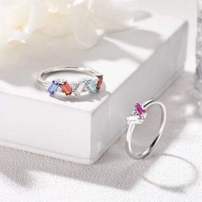 A custom baguette birthstone ring with multiple colored stones set in a white gold band, displayed on a white surface with a floral background.