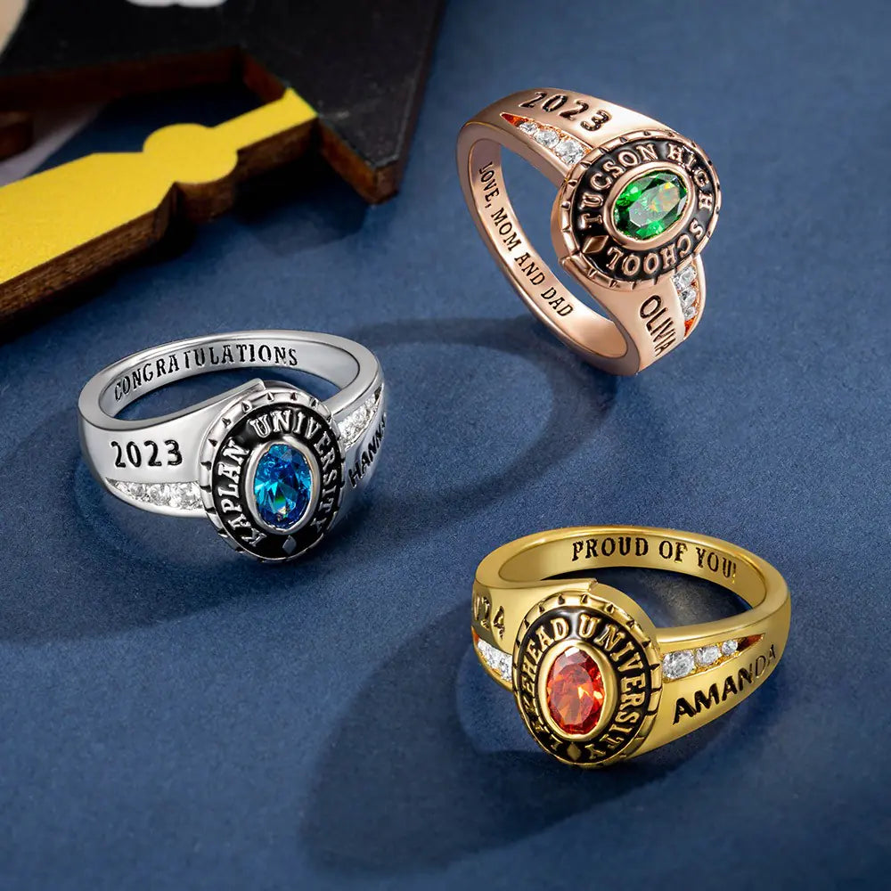Three custom class rings for women, made of brass or sterling silver, with engraved text and personalized birthstones, displayed on a blue surface with a shadow cast over them.