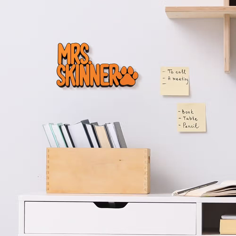 Orange 3D wall decal reading 'MRS. SKINNER' with a paw print, mounted on a white wall above a wooden organizer box in a styled home office setting.