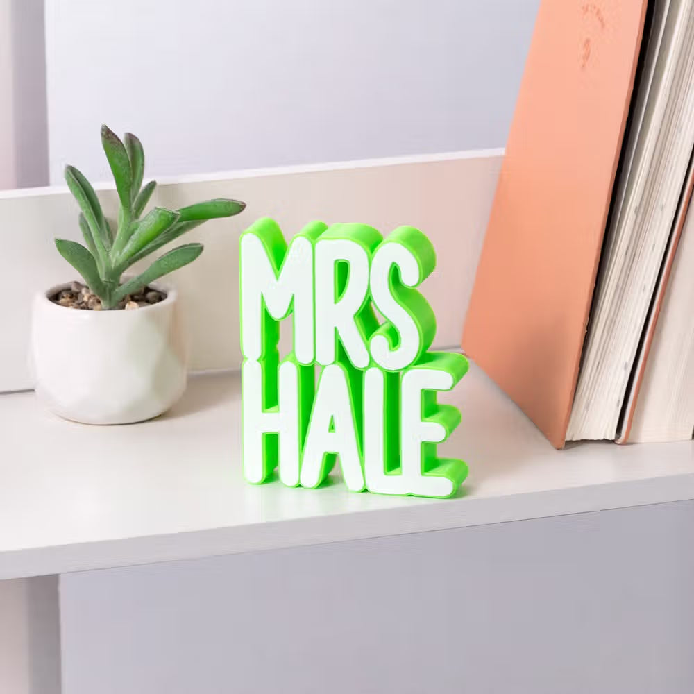 Green personalized 3D teacher name sign displayed on a classroom shelf with books and a plant