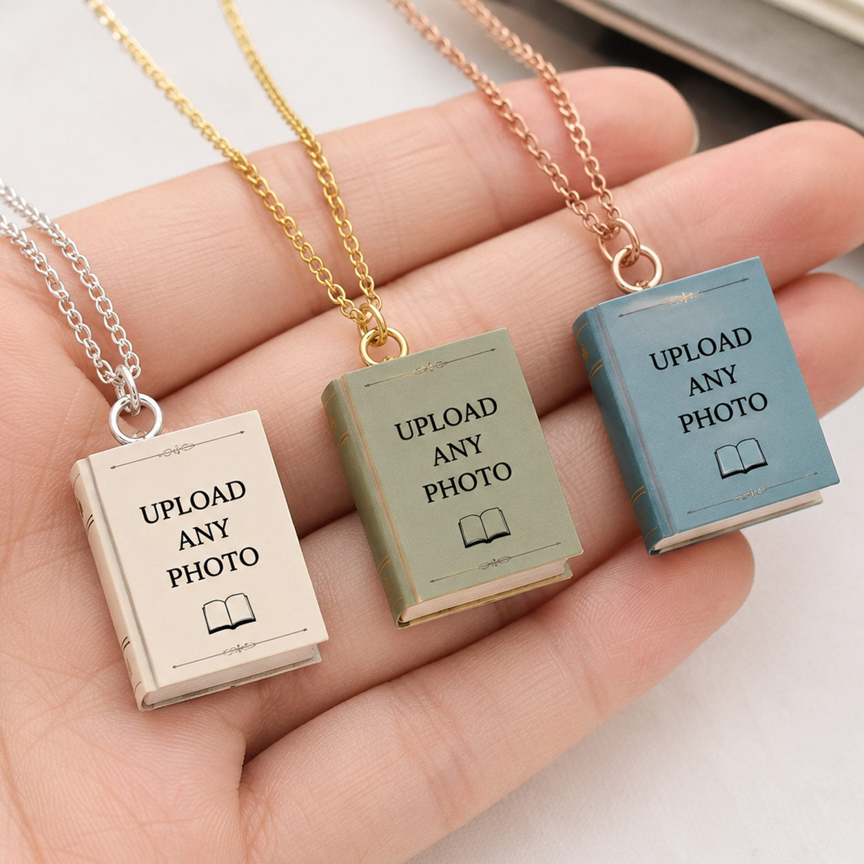 Hand holding three personalized miniature book cover necklaces with silver, gold, and rose gold chains, featuring cream, sage, and blue book covers with "Upload Any Photo" sample designs.