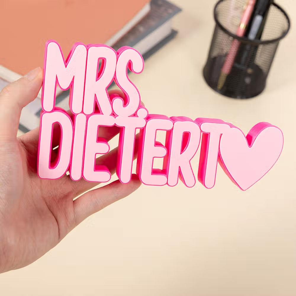A hand holds a pink custom 3D decorative sign spelling 'MRS. DIETERT' with a heart at the end, set against a softly blurred desk background.